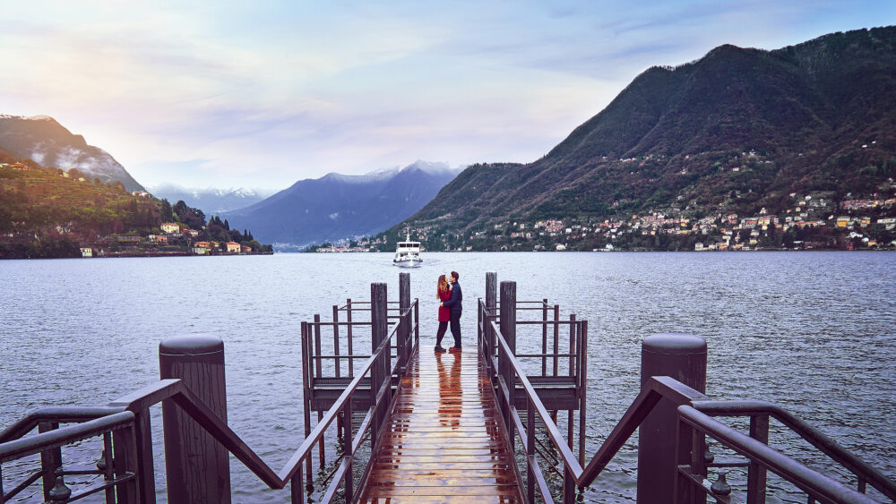 A couple embraces on a dock over a scenic lake, with soft lighting and mountains framing the background.