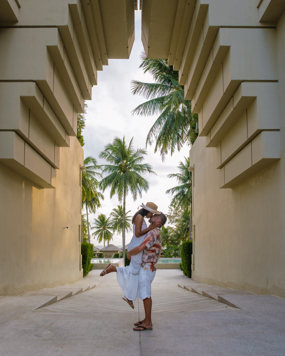 A man lifts a woman into a kiss beneath modern architectural structures, framed by palm trees and golden sunlight.