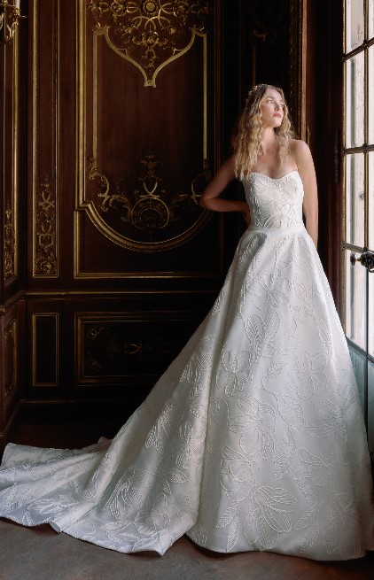 A bride stands beside a tall window wearing a voluminous ballgown with a textured floral pattern and strapless neckline.