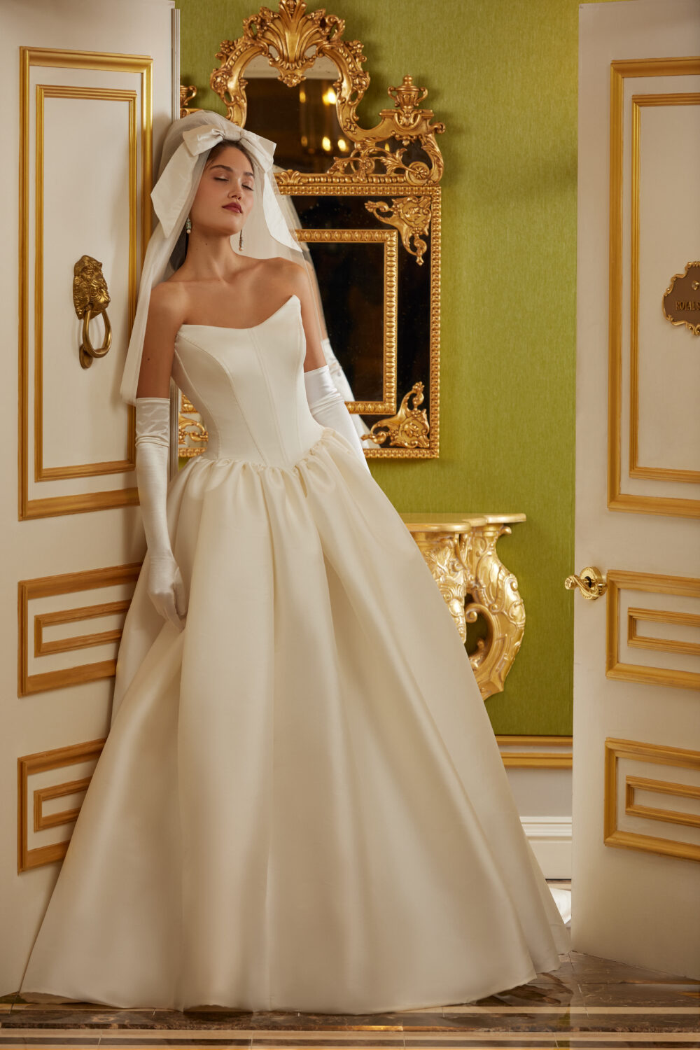 A bride poses beside an opulent gold-trimmed doorway wearing a full ballgown with gloves and a statement bow headpiece.