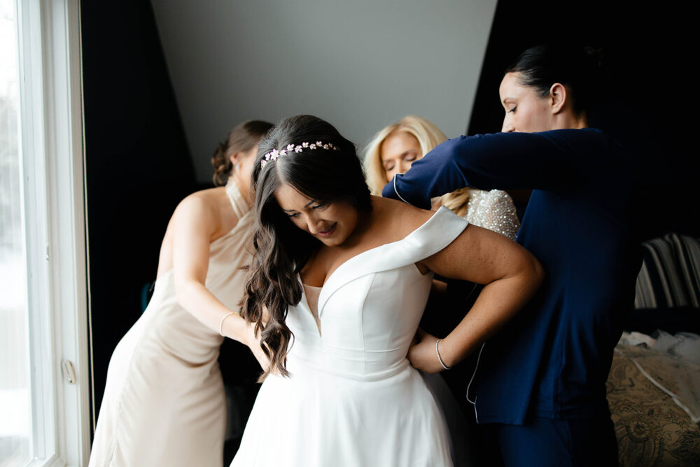 Bride in off-the-shoulder gown surrounded by bridesmaids helping her get dressed.