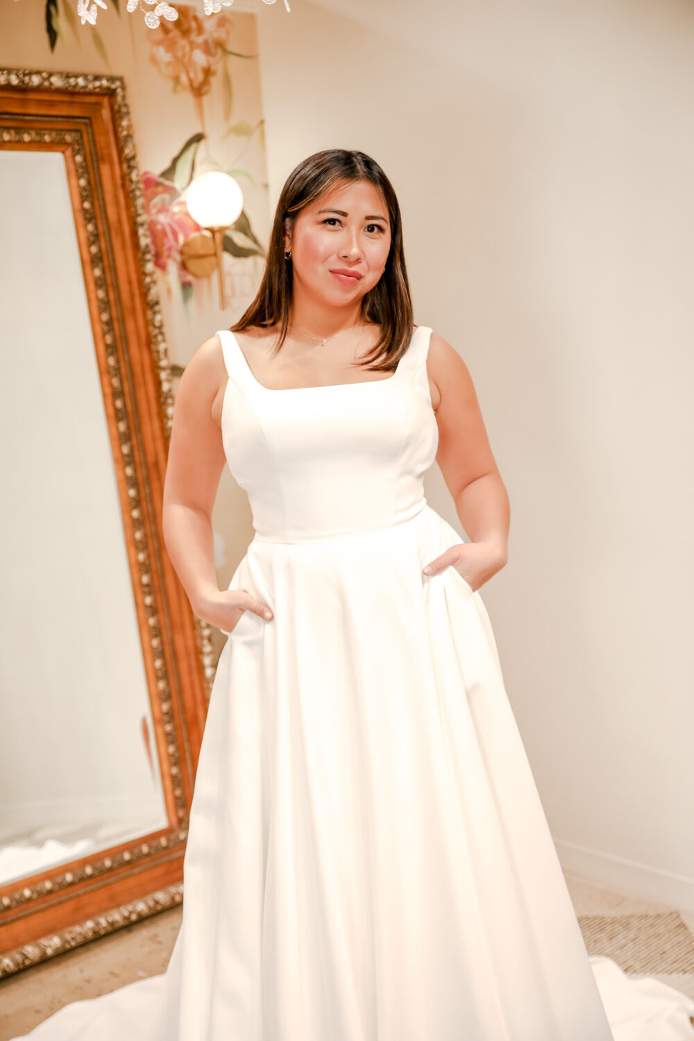 A bride stands in front of a vintage mirror wearing a satin gown with a structured square neckline and deep pockets, surrounded by warm lighting and floral wallpaper.