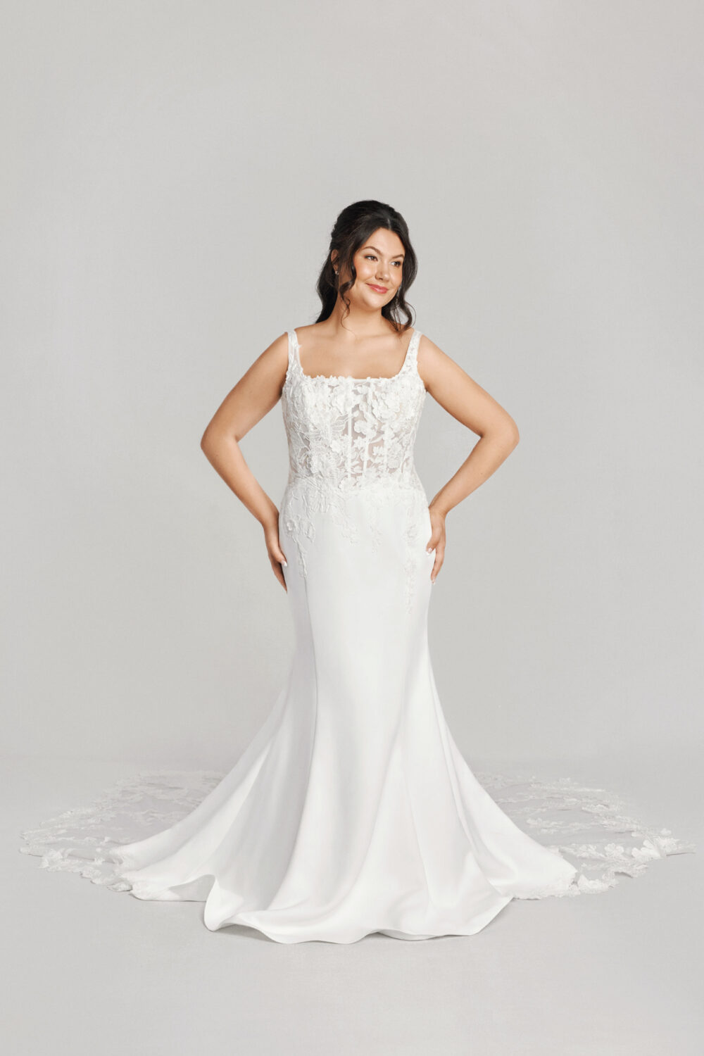 A bride poses confidently in a fitted gown featuring a lace bodice with floral appliqué and a sleek satin skirt that flows into a chapel-length train.