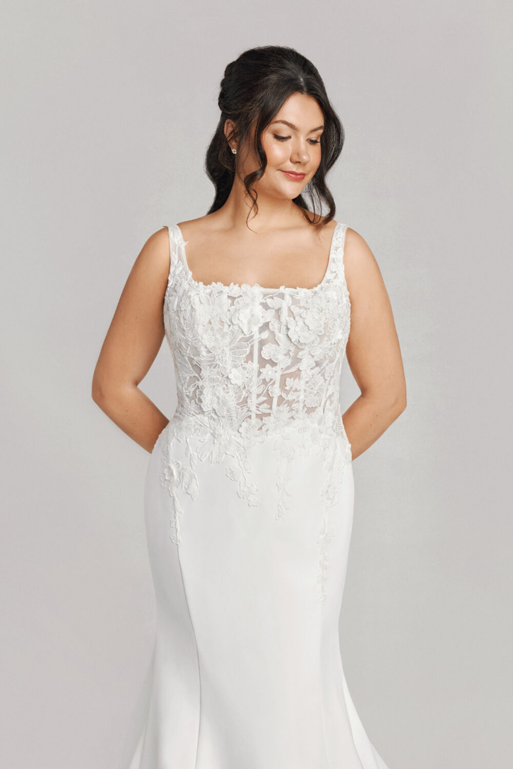 The bride gently clasps her hands behind her back, highlighting the intricate lace detailing on the structured bodice of her square neckline gown.