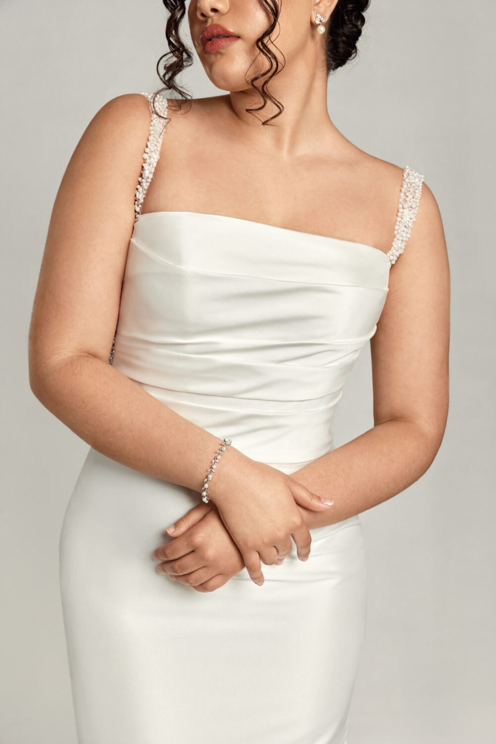 A close-up of a bride’s ruched satin gown shows her crossed arms and delicate bracelet, along with beaded straps and a straight neckline.