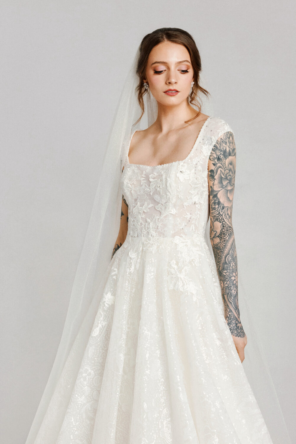 A bride with tattooed arms gazes down in a romantic lace gown with a sheer bodice, square neckline, and full skirt under a glittering veil.