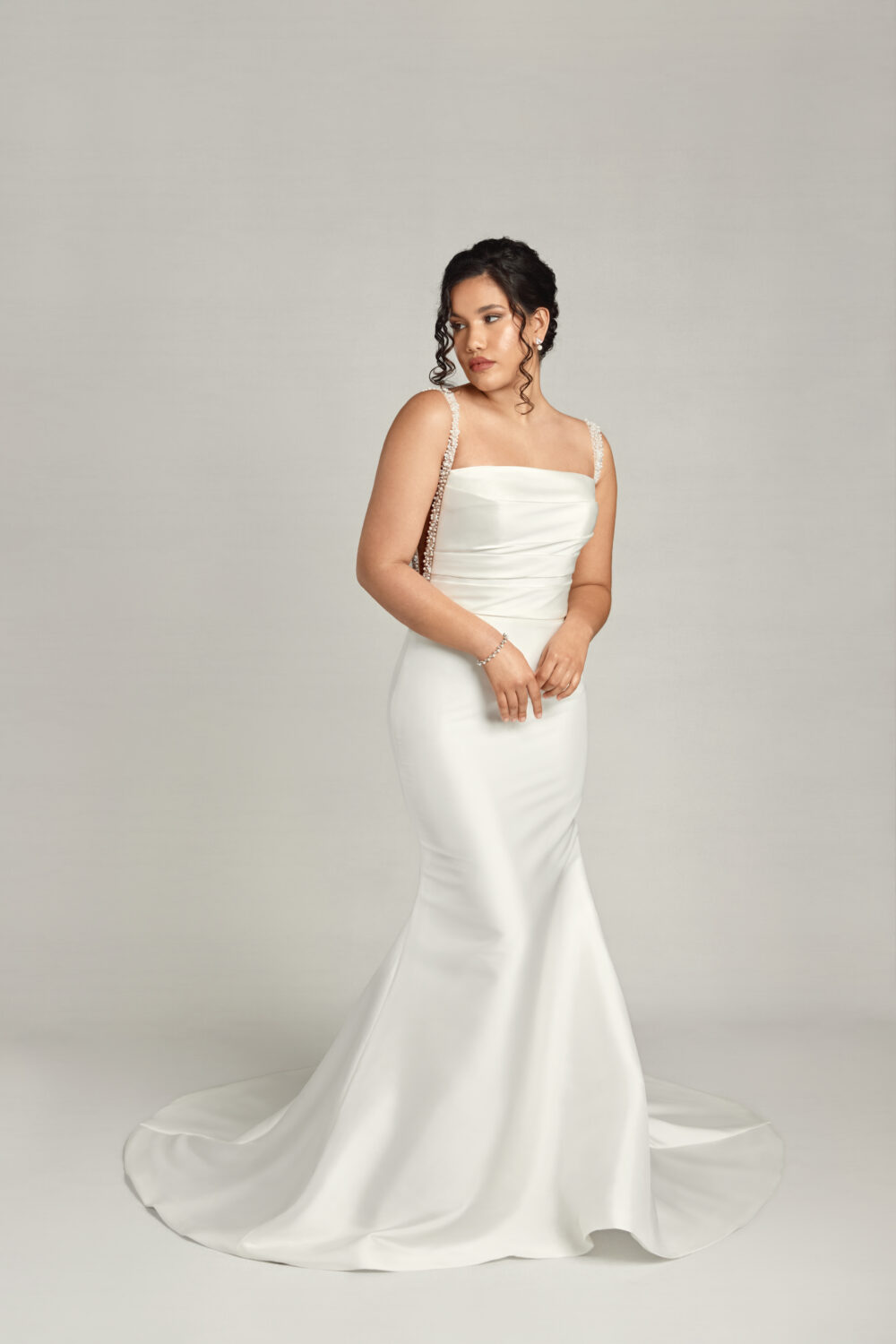 The bride looks off to the side in a full-length portrait, wearing a satin gown with a straight neckline and beaded straps over a flared skirt.