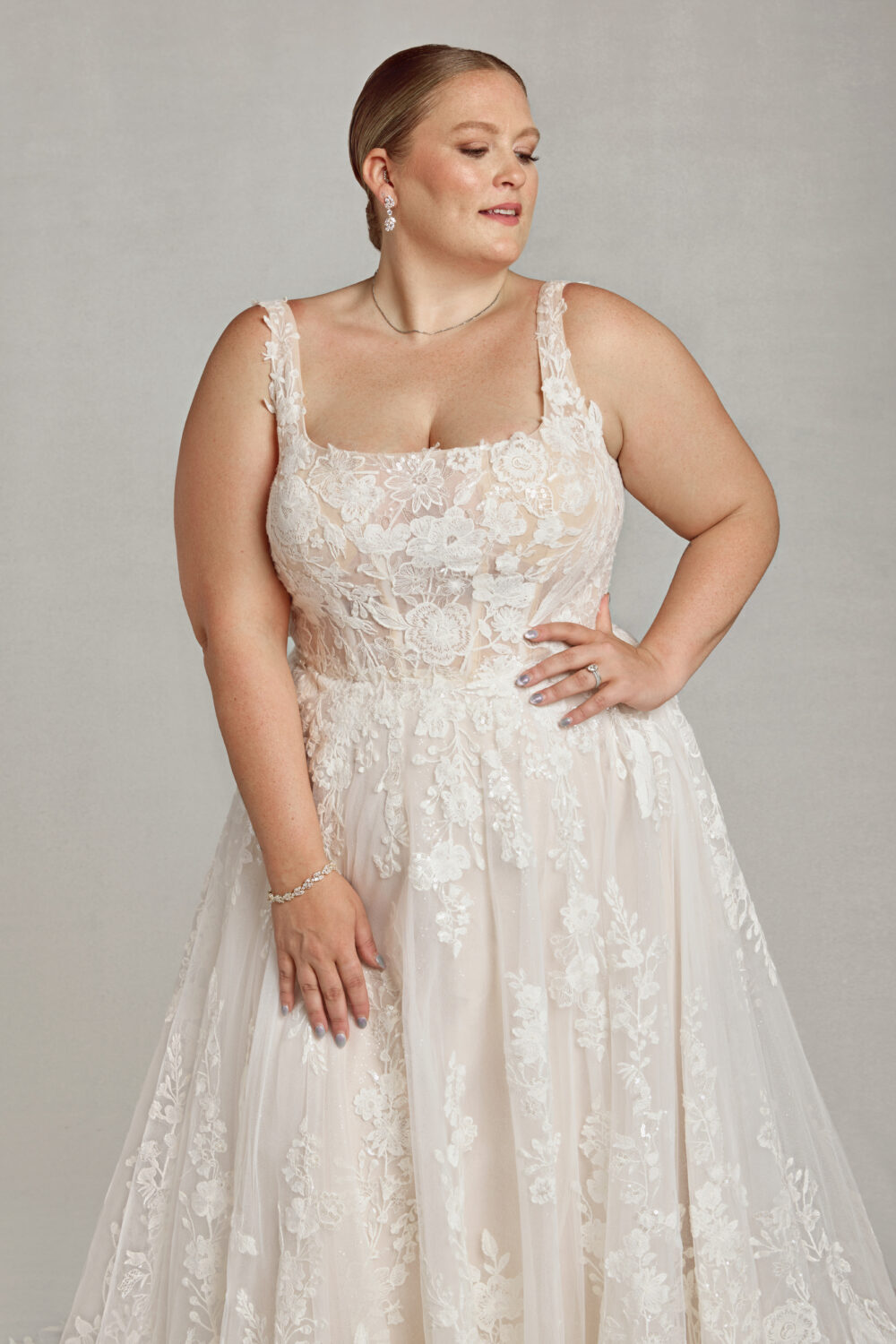A bride with her hair in a sleek bun poses in a romantic A-line gown featuring thick straps, a floral lace bodice, and a softly flowing skirt.