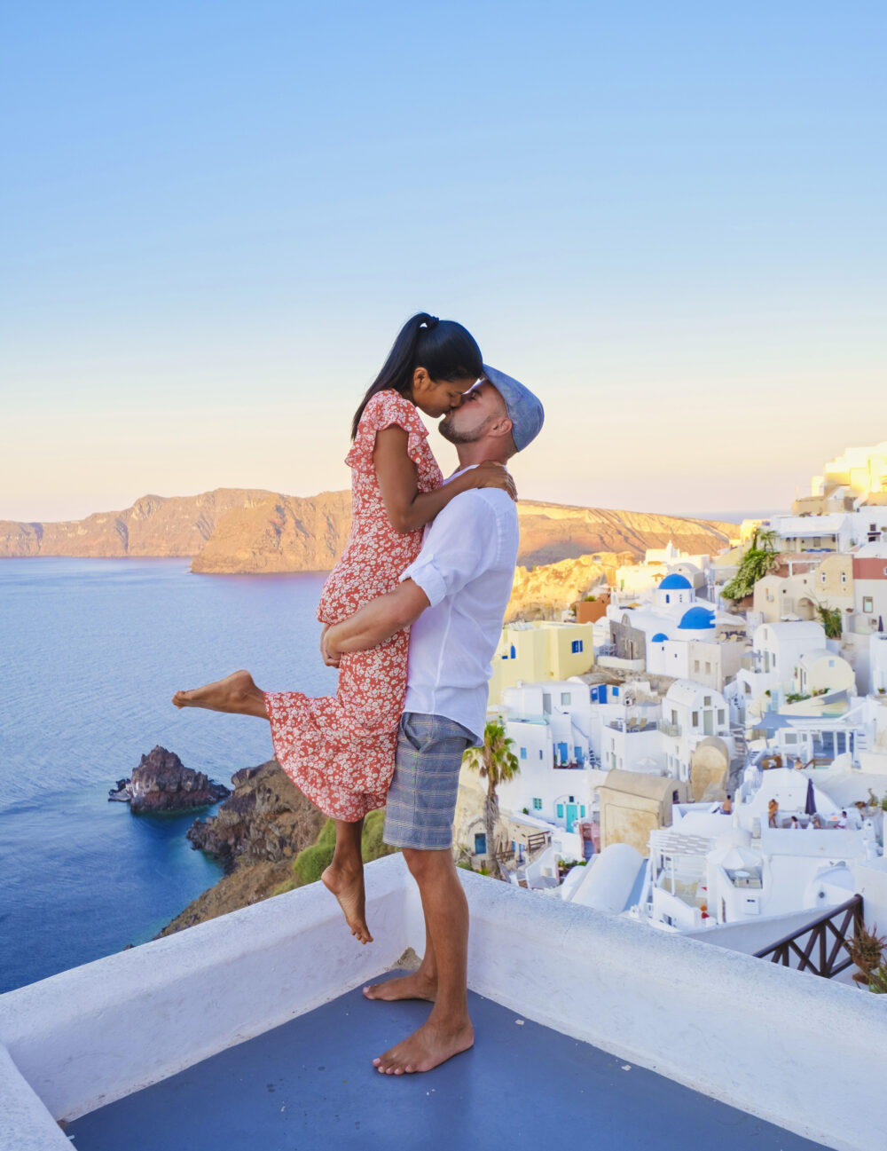 A barefoot couple kisses on a rooftop at sunset, with the white buildings and blue domes of Santorini visible behind them.