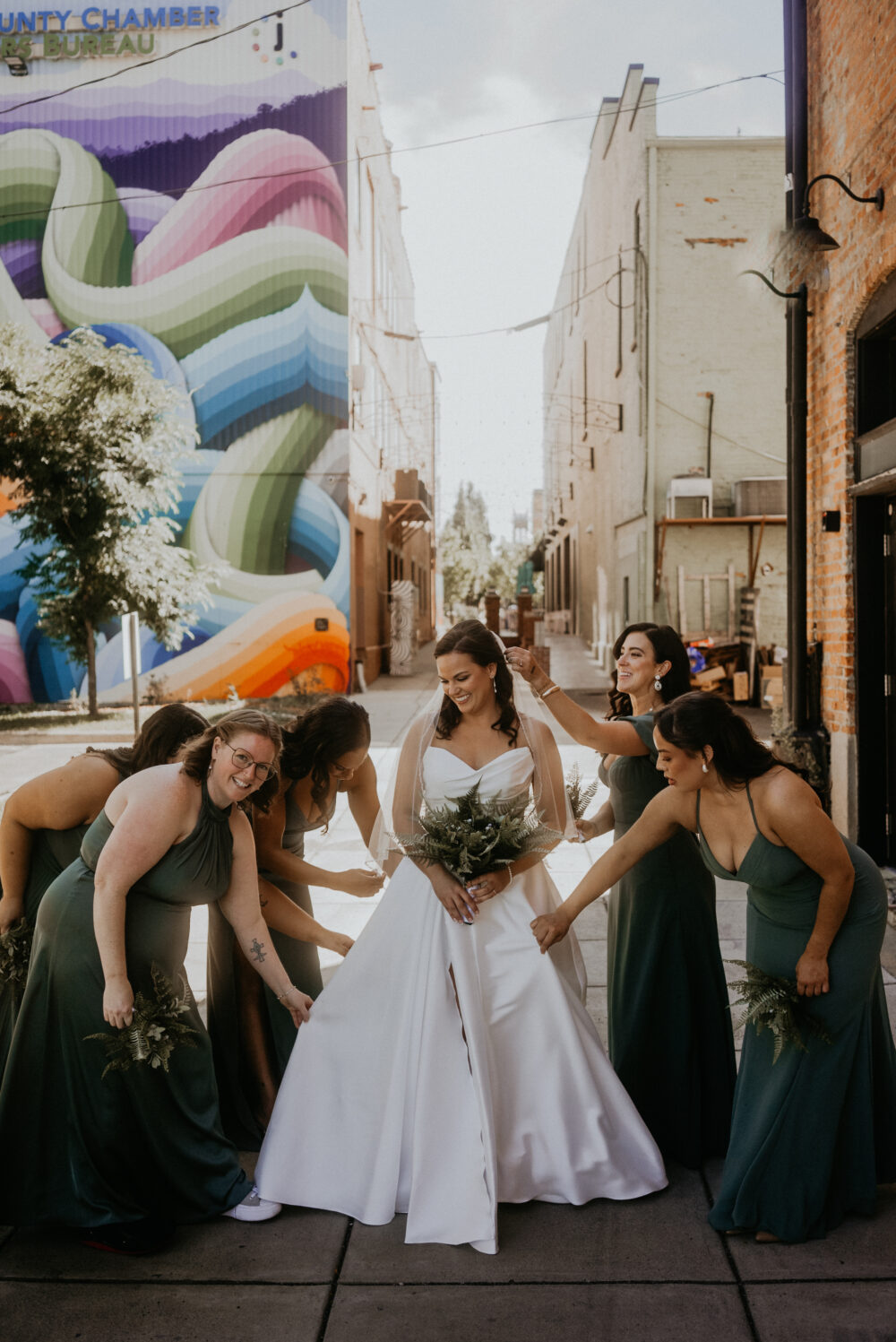 A bride stands in an alleyway with a group of bridesmaids adjusting her strapless gown, with a vibrant mural in the background.