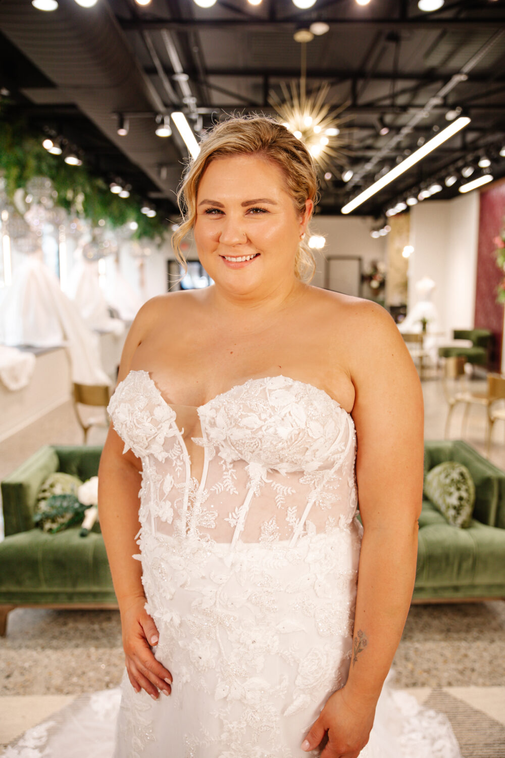 A smiling bride tries on a strapless lace wedding gown with a plunging neckline and sheer corset bodice at a bridal boutique.