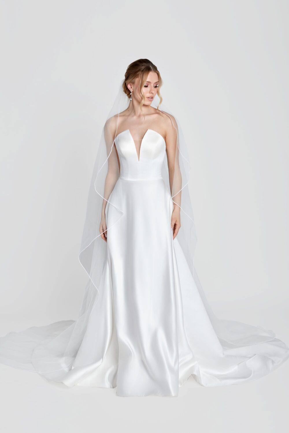 Bride wearing a minimalist strapless wedding gown with a plunging neckline and sheer veil, posed in a bright studio.