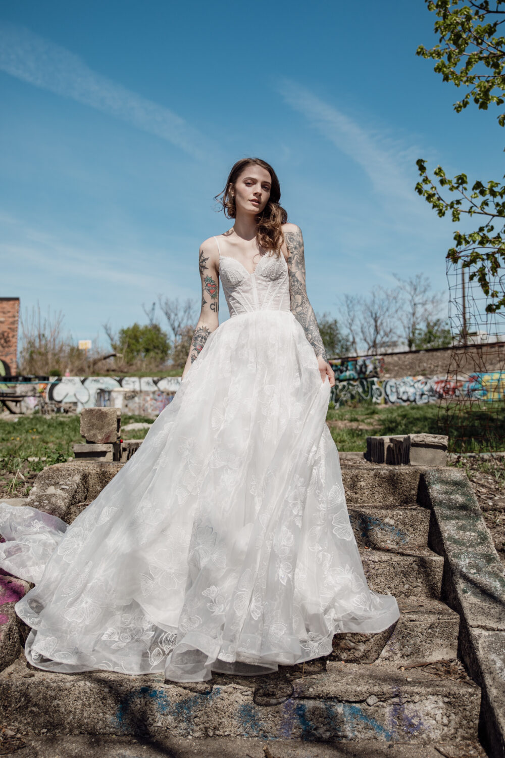 A bride with tattoos poses outdoors on stone steps in a lace corset wedding dress with spaghetti straps and a full floral-embroidered skirt.