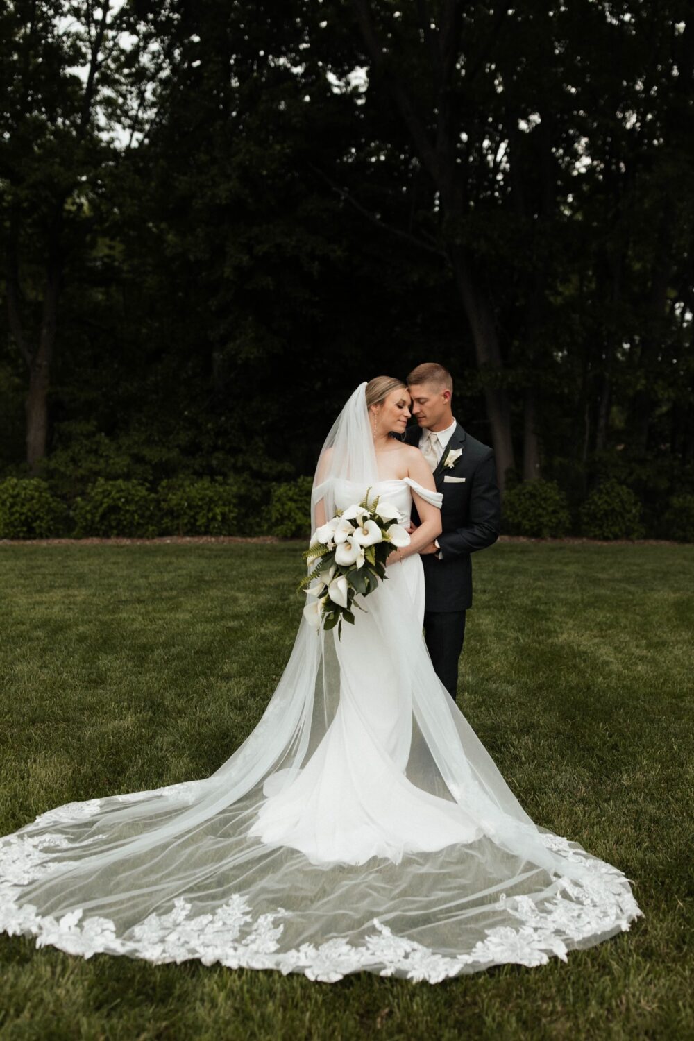 A bride with a long cathedral veil and bouquet of white calla lilies poses with her groom on a grassy lawn surrounded by trees.