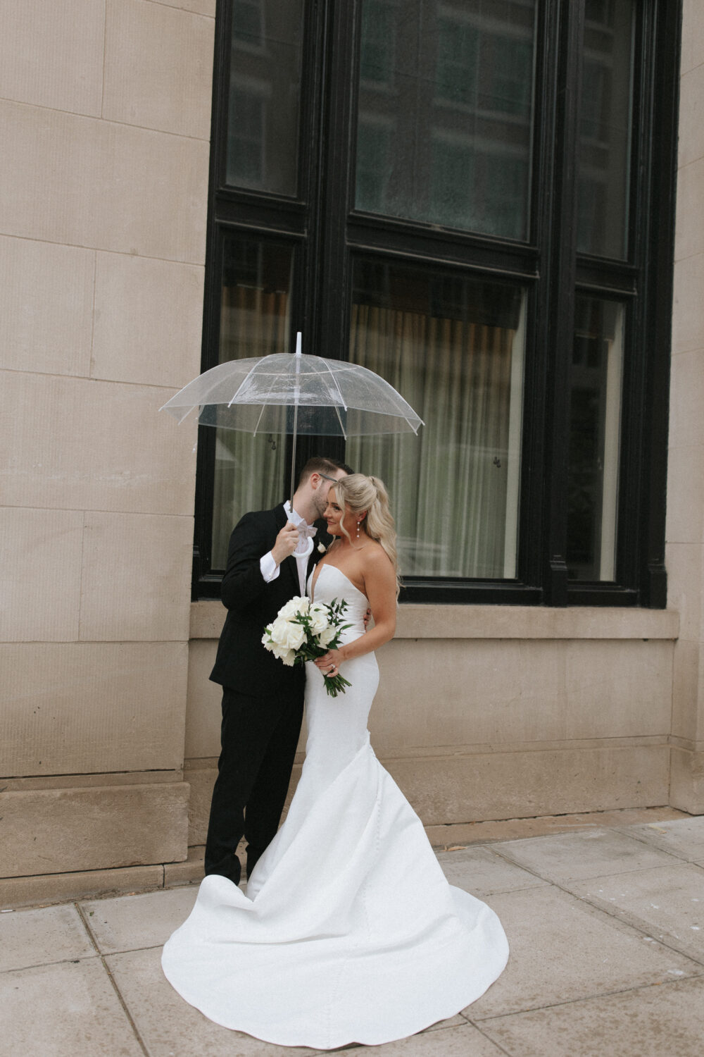 A bride in a strapless gown shares a romantic moment with her groom under a clear umbrella in front of a downtown building.