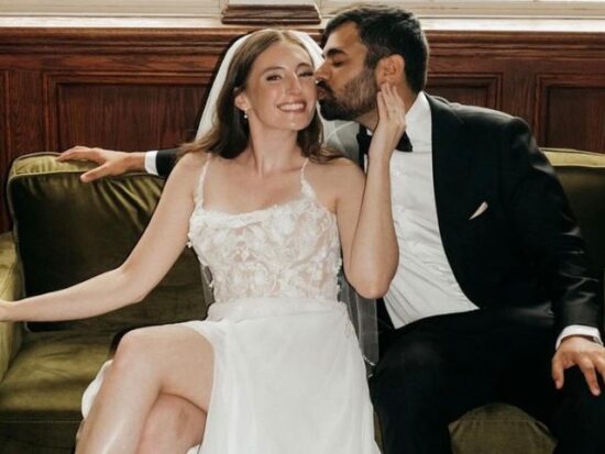 A cropped view of a bride and groom sitting on a green couch, with the groom kissing the bride’s cheek while she smiles.