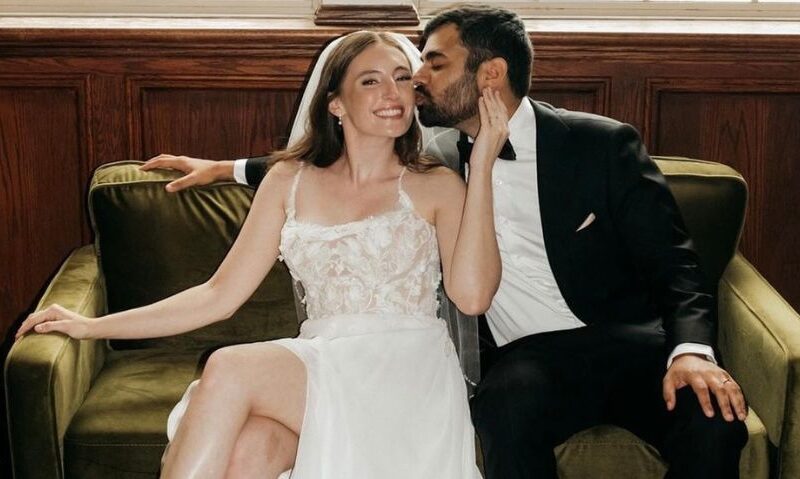 A cropped view of a bride and groom sitting on a green couch, with the groom kissing the bride’s cheek while she smiles.