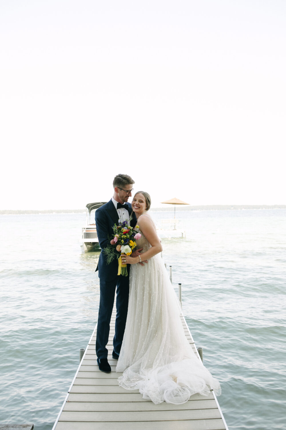 A bride in a beaded A-line gown holds a bouquet and laughs while her groom wraps his arms around her on a dock by the water.
