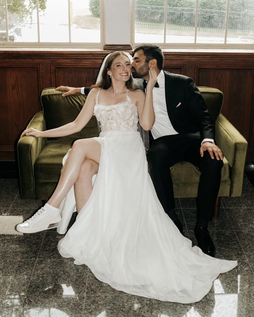 A bride in a lace wedding dress with a thigh-high slit and sneakers sits on a green velvet couch while her groom kisses her cheek.