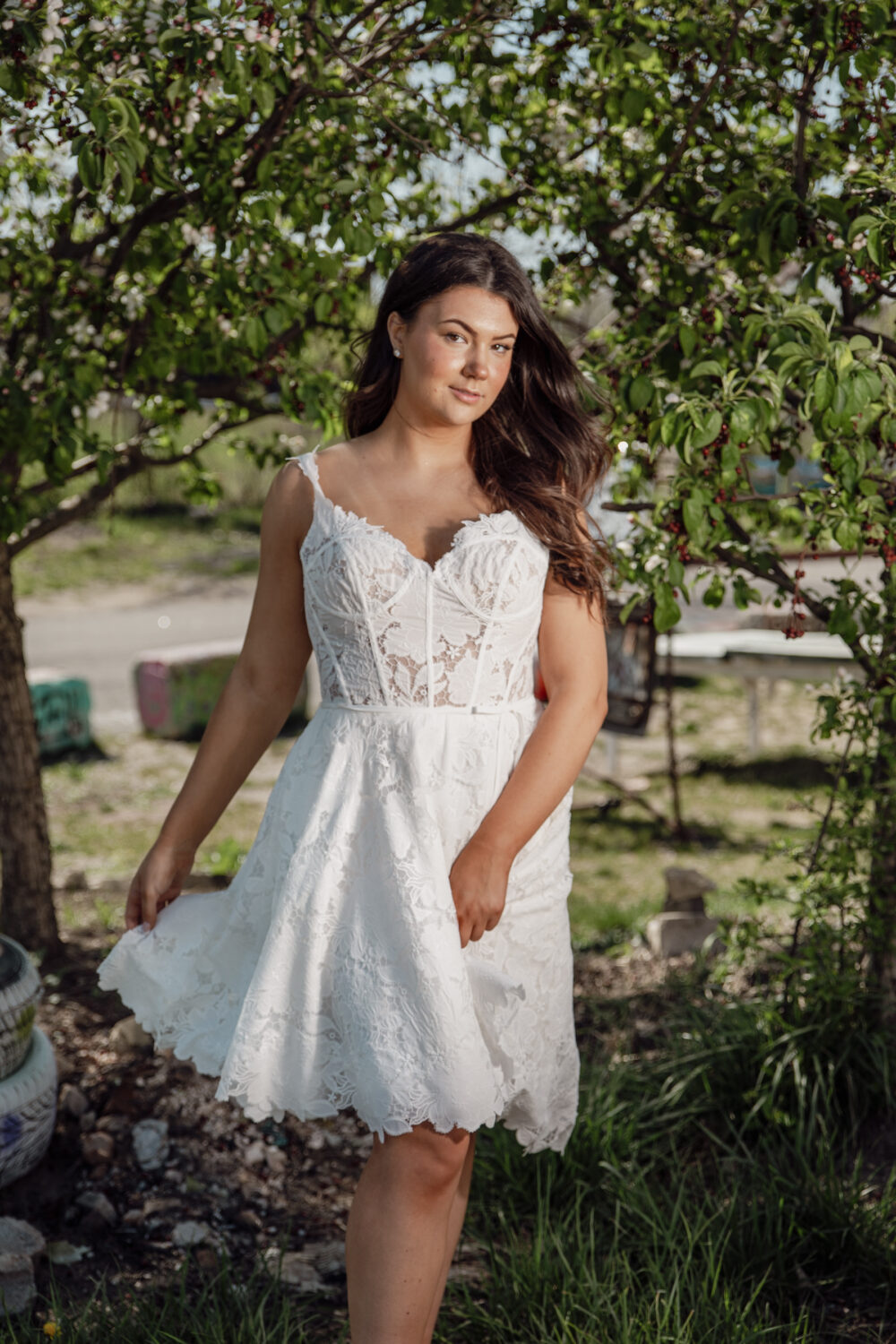 A bride poses outdoors in a short lace wedding dress with spaghetti straps and a semi-sheer corset bodice surrounded by leafy green trees.