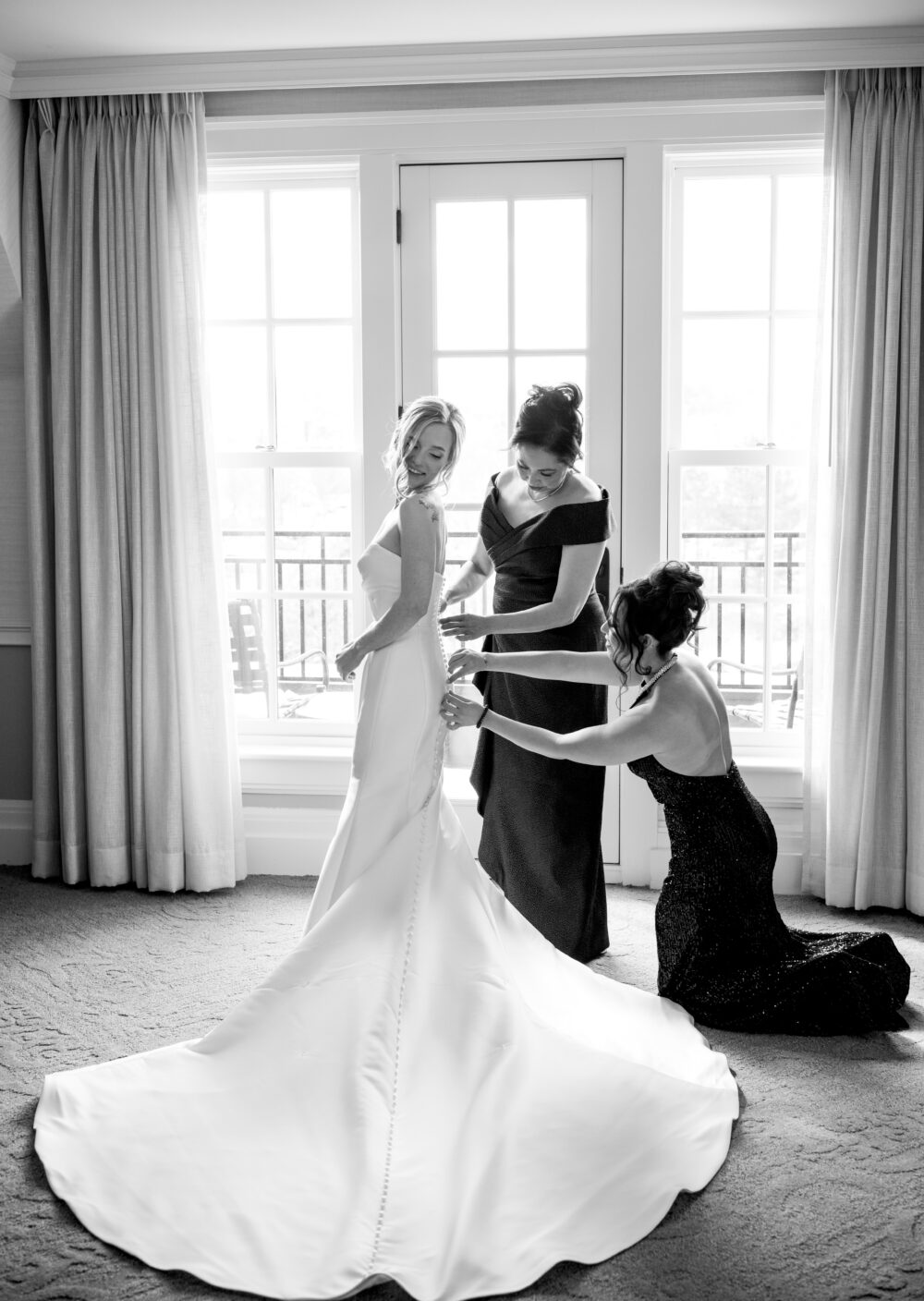 Black-and-white image of bride smiling as bridesmaids fasten her gown in front of window.