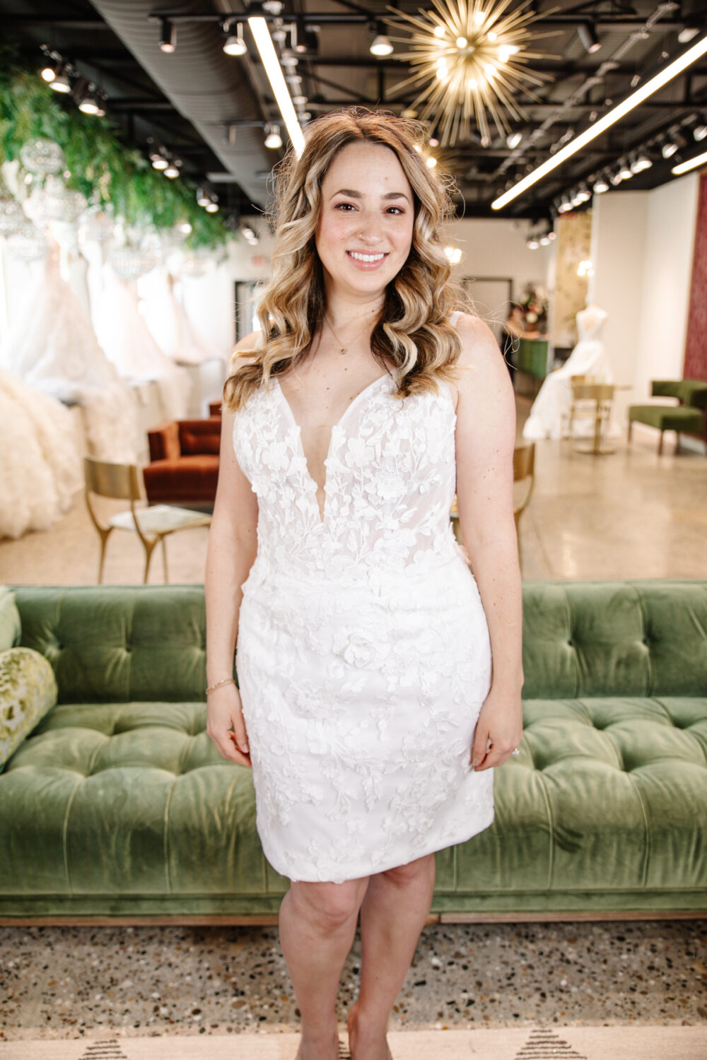 A bride smiles warmly inside The Wedding Shoppe while wearing a floral lace fitted wedding dress with a plunging neckline and sheer bodice.