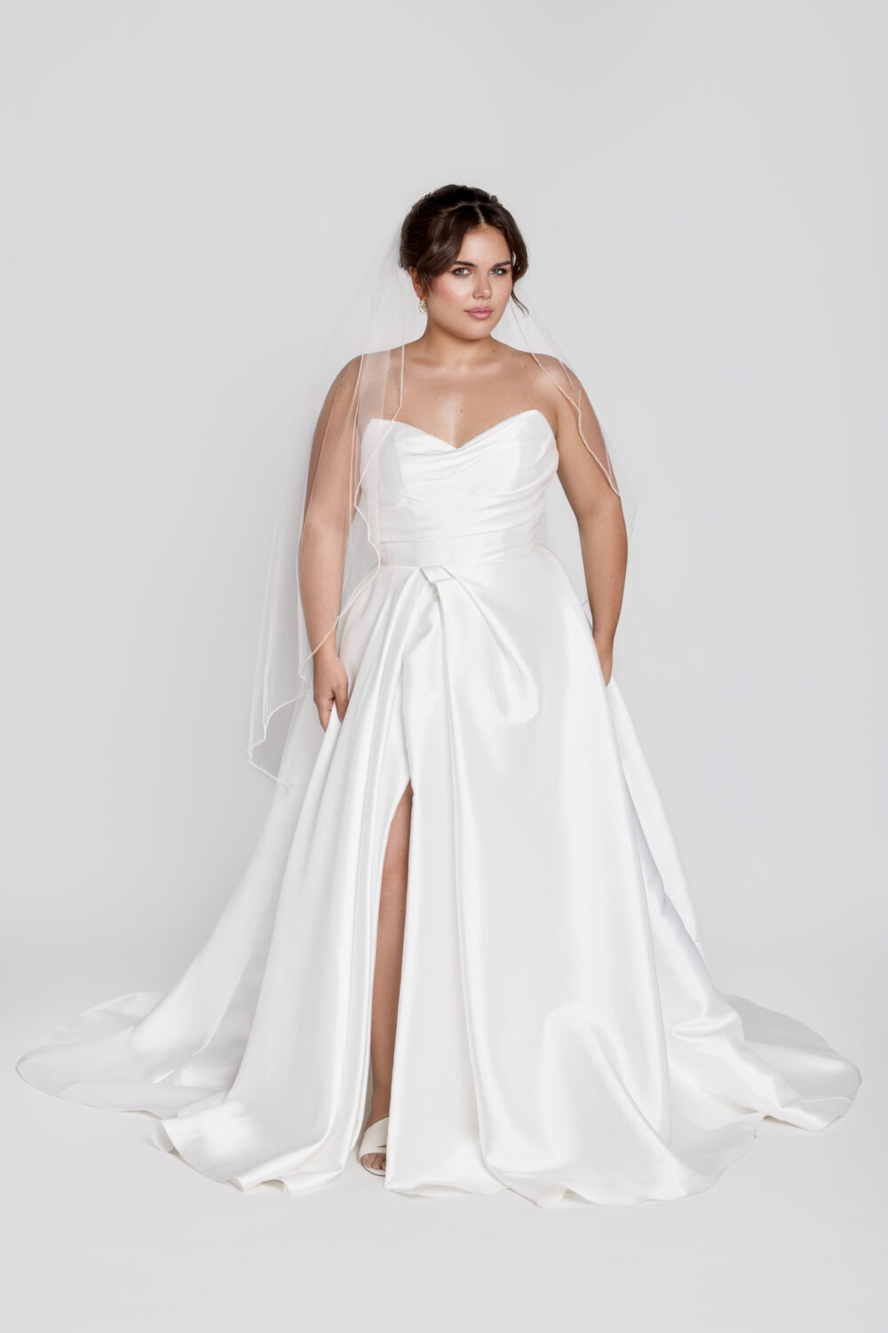 A bride wears a strapless A-line wedding gown with ruching and a front slit, styled with a fingertip-length veil and neutral heels.