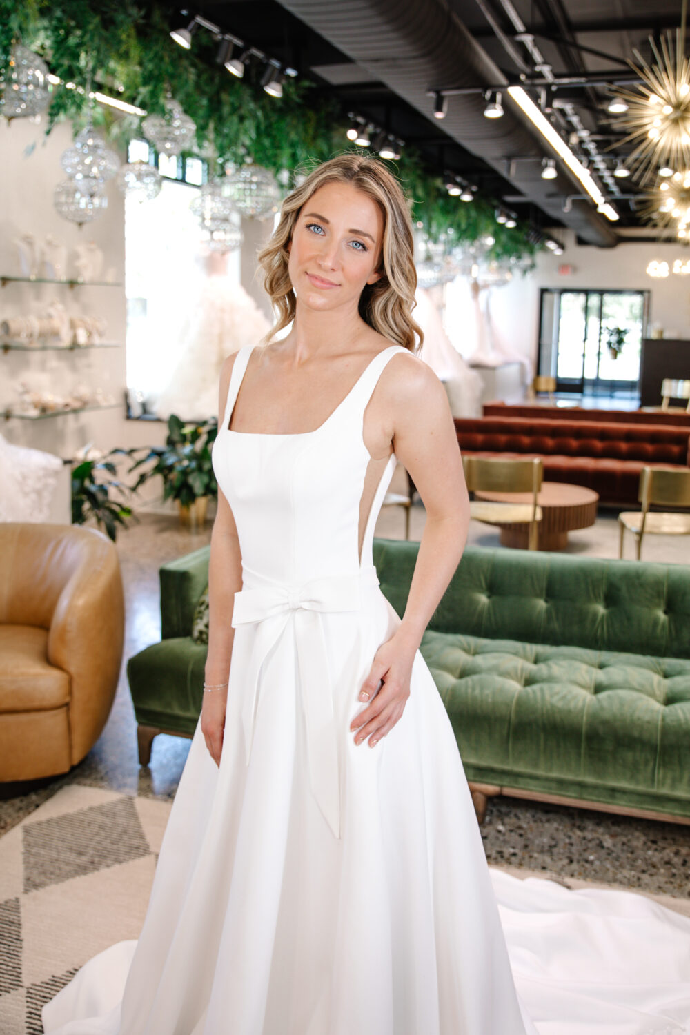 A bride models a minimalist square-neck A-line wedding dress with a flat bow at the waist in a chic bridal showroom.