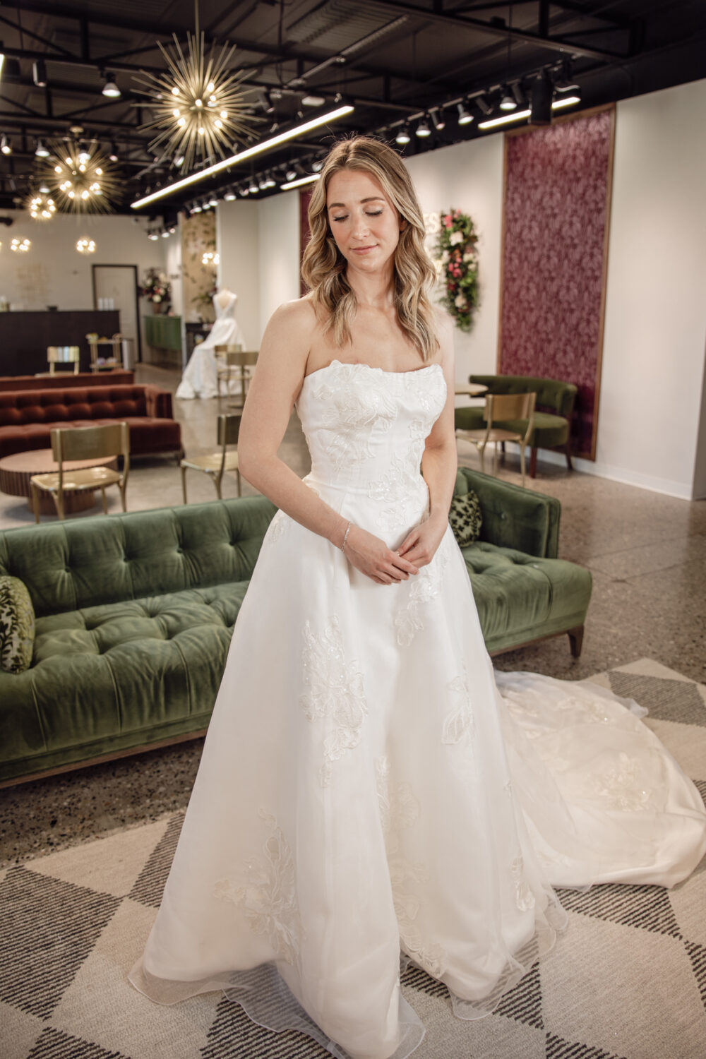 A bride wears a strapless embroidered A-line wedding dress with a train inside a modern bridal boutique with green velvet furniture.