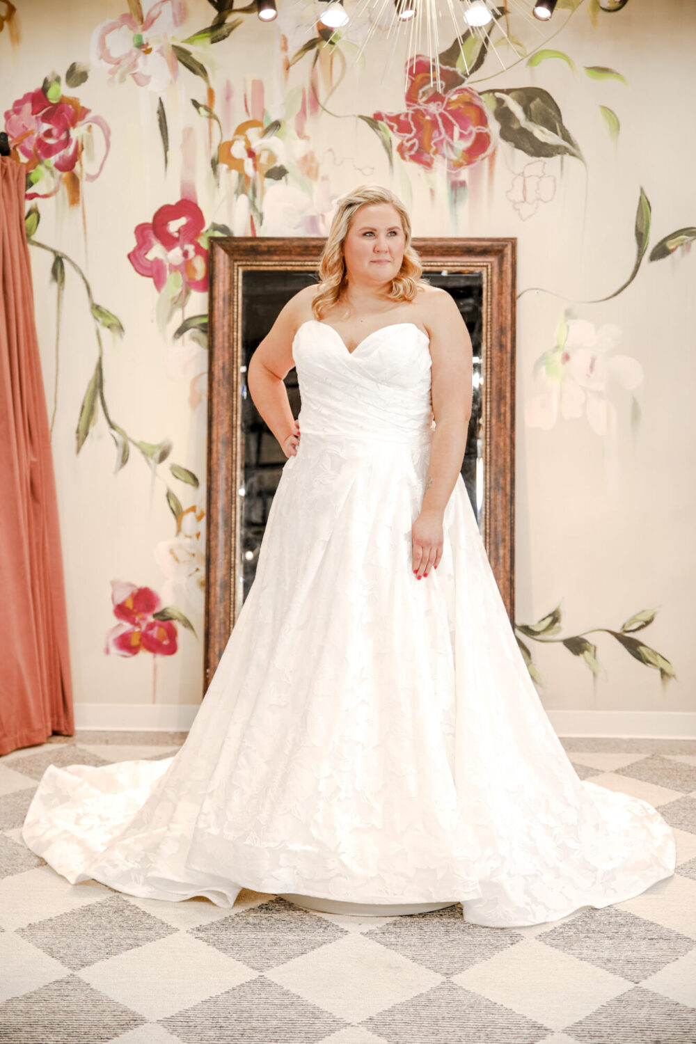 Bride in a strapless floral jacquard gown poses in front of a mirror and a painted floral wall in a boutique fitting room.