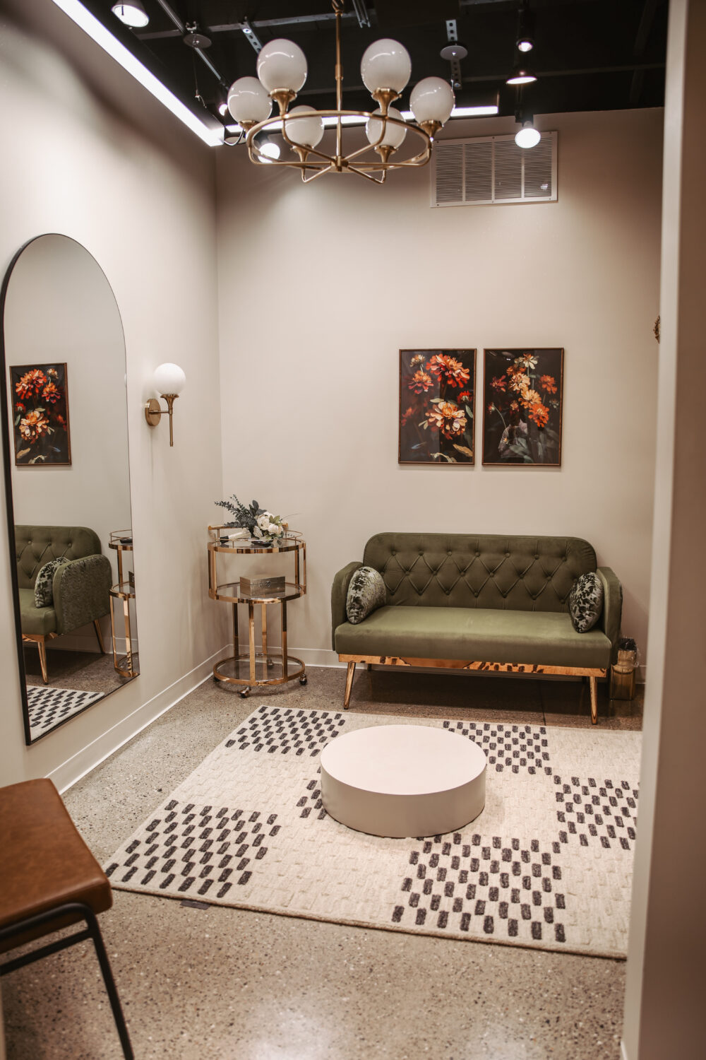 A stylish bridal suite features an olive green tufted couch, round gold bar cart, and warm lighting, creating a cozy pre-appointment space.