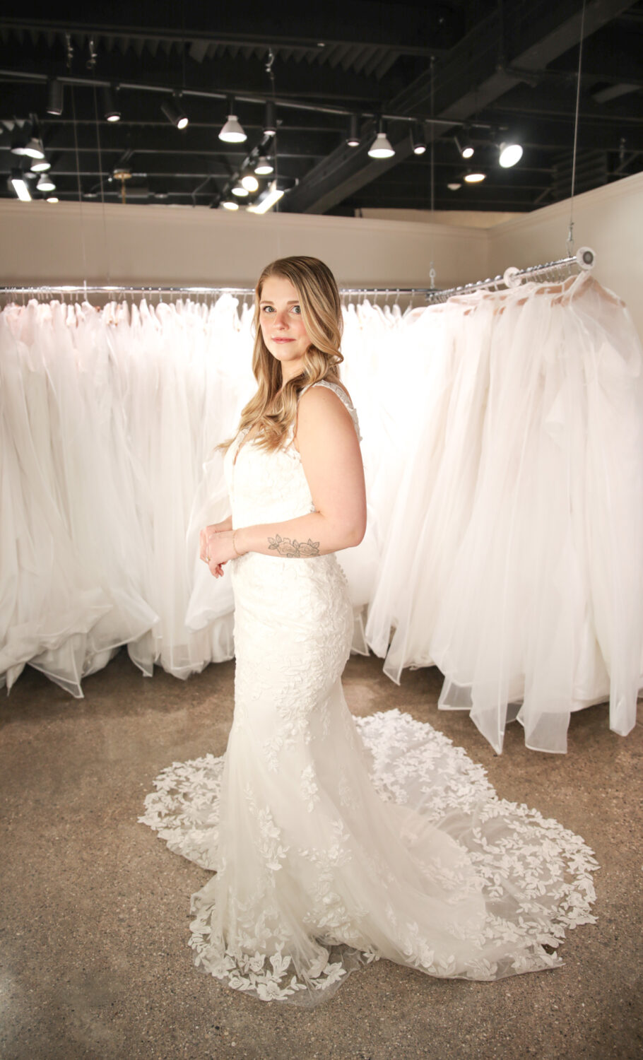 A bride stands confidently in front of a wall of wedding dresses wearing a fitted lace gown with a floral lace train and sheer straps.