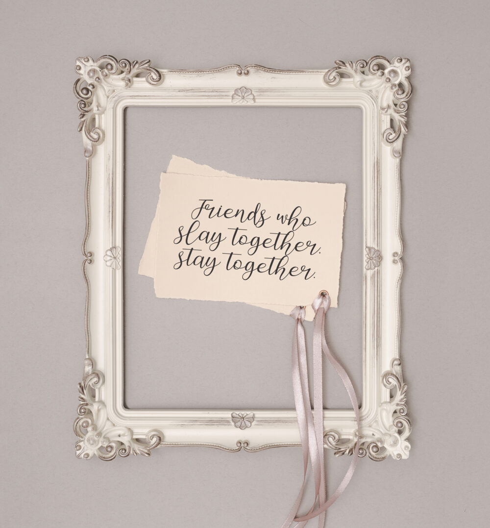 A vintage-style white picture frame holds a torn card that reads “Friends who slay together, stay together,” with pink satin ribbons hanging from the bottom.
