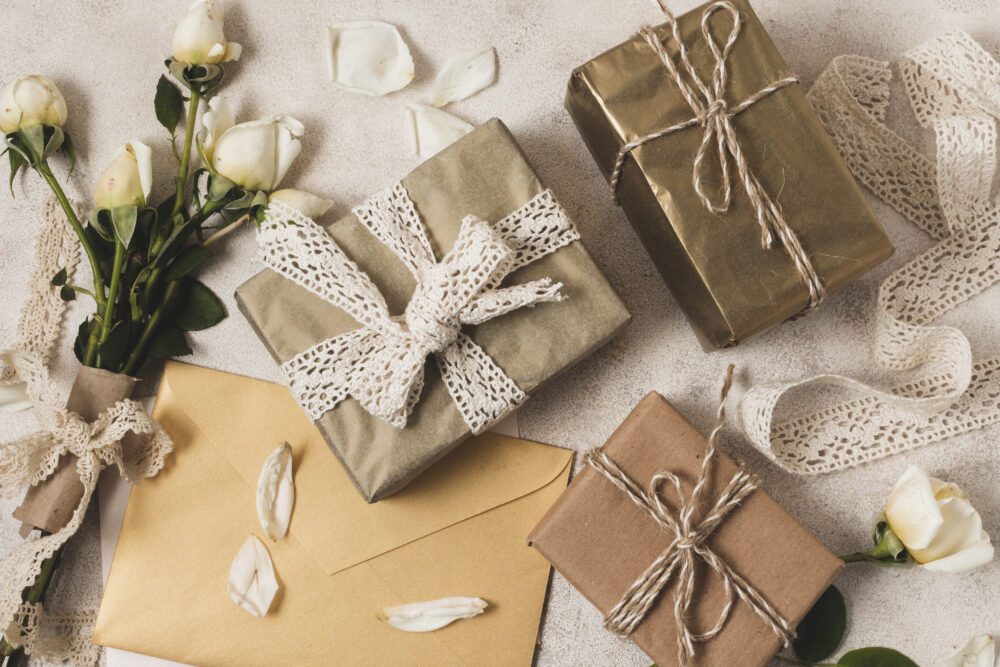 A flat lay of three beautifully wrapped gifts with lace and twine ribbons, surrounded by white rosebuds, golden envelopes, and scattered flower petals.