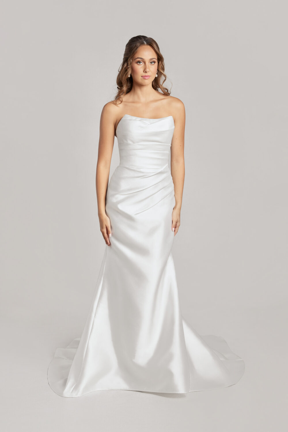 A model poses against a light background in a sleek satin wedding dress with subtle ruching and a straight neckline.
