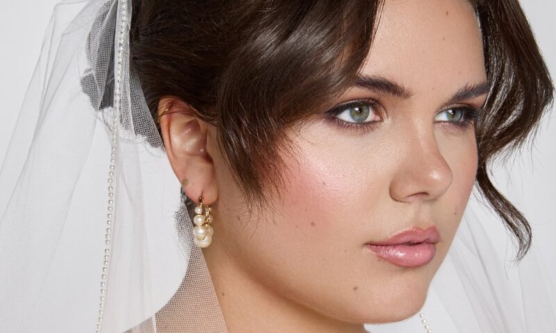 A bride wears a pearl-trimmed wedding veil, paired with drop pearl earrings and soft romantic makeup.