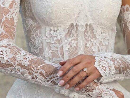 A bride poses in front of a textured wall, wearing a structured white ball gown with a square neckline and wide straps, holding orchids.