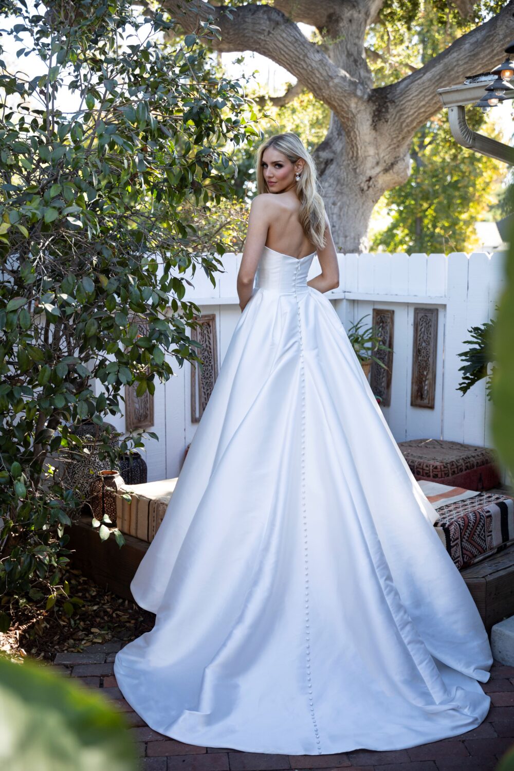 A bride in a strapless satin ball gown stands in a garden setting, showing off a row of covered buttons trailing down the back.