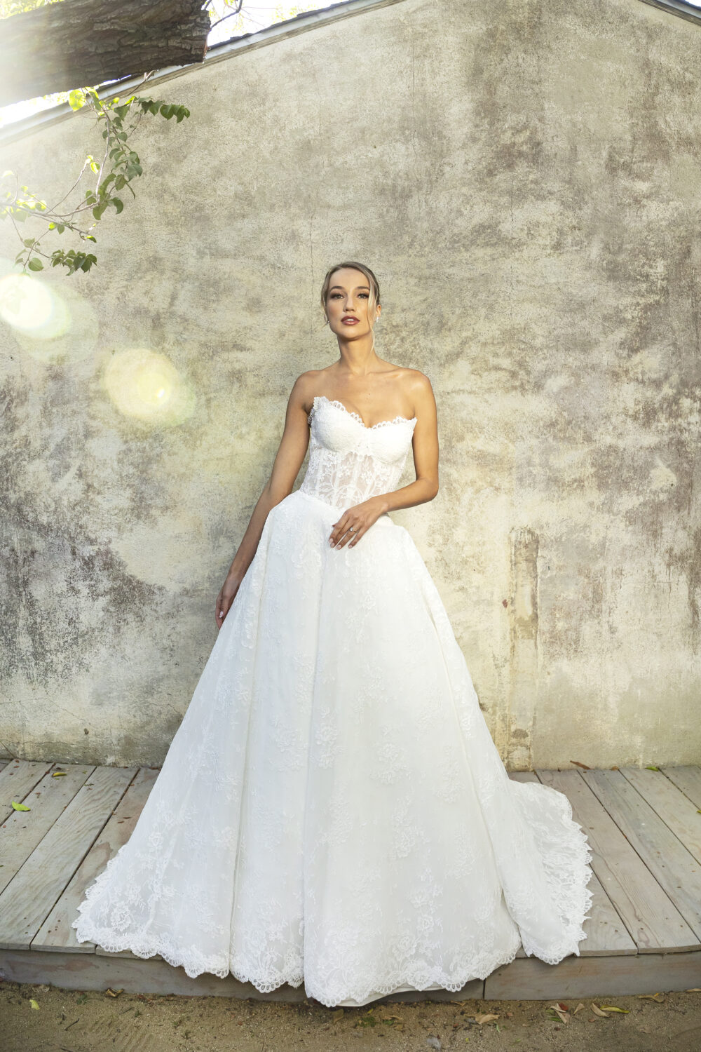 A bride poses against a rustic wall, wearing a strapless lace gown with a scalloped neckline and a soft, flowing A-line skirt.