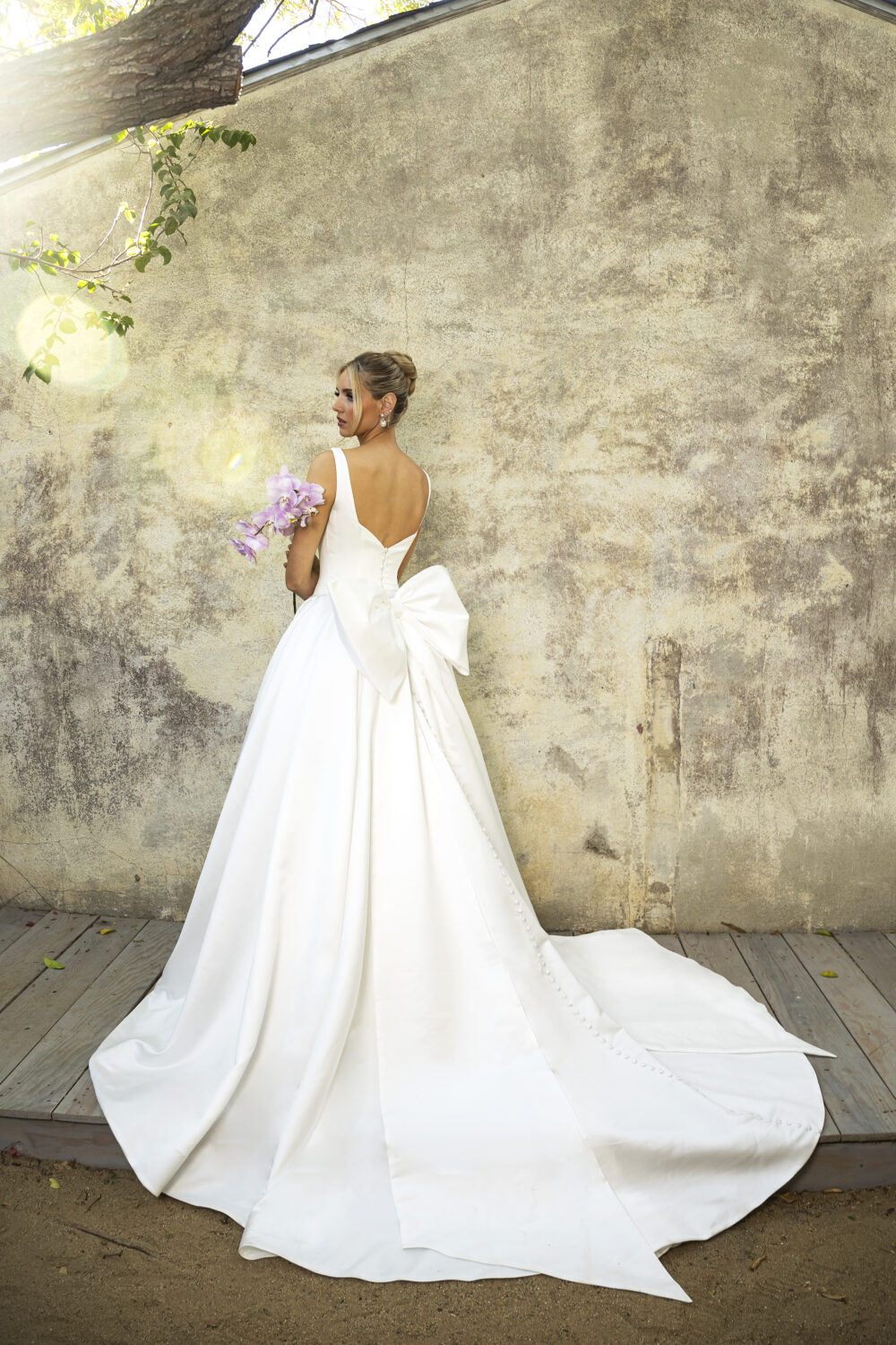 A bride stands outdoors holding light purple orchids, wearing a white ball gown with a dramatic back bow and a V-back neckline.