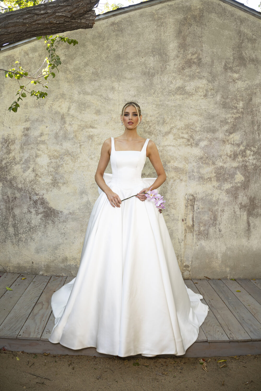 A bride poses in front of a textured wall, wearing a structured white ball gown with a square neckline and wide straps, holding orchids.