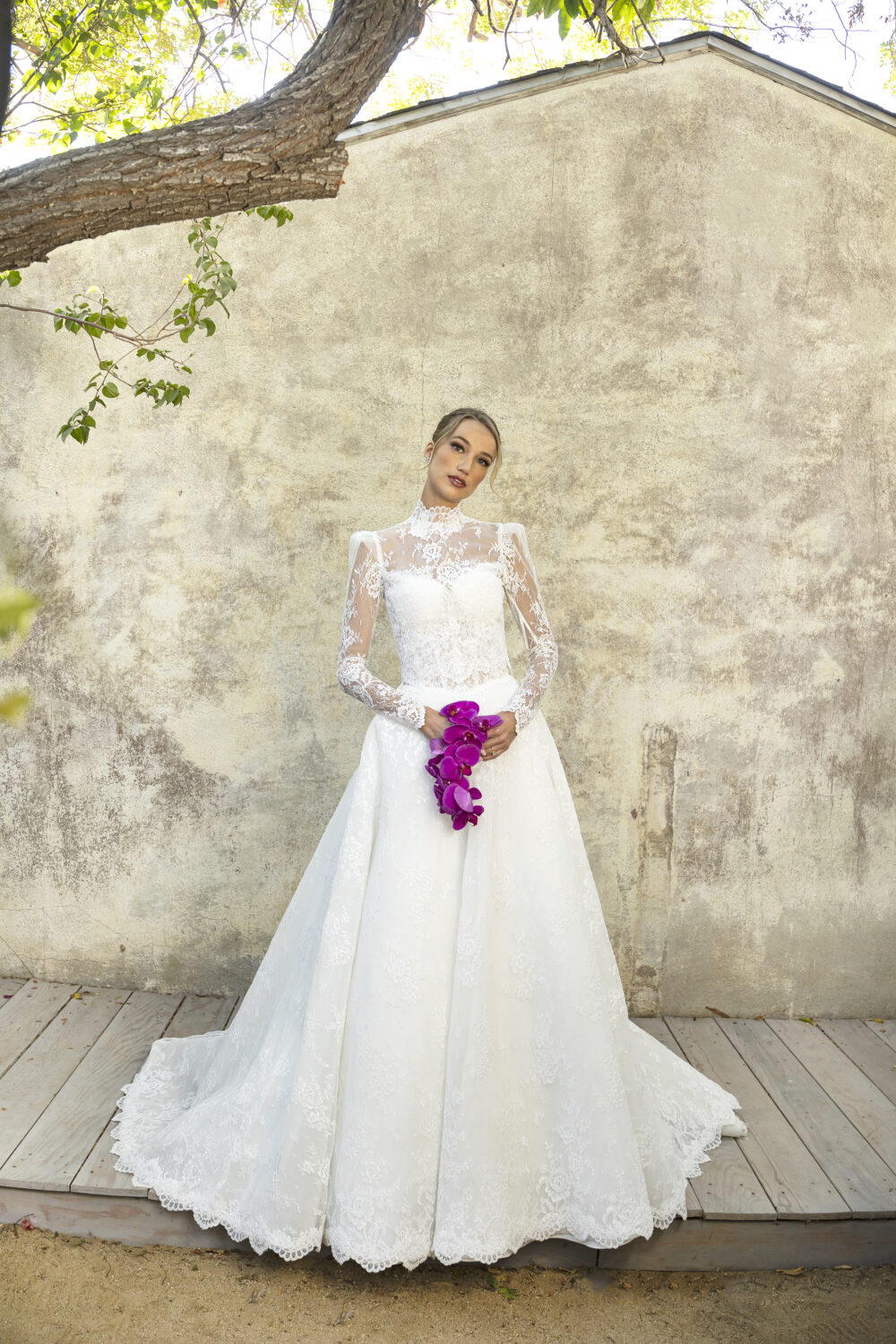 A bride poses against a textured wall in a long sleeve lace wedding gown with a mock neck and full ball gown skirt, holding purple flowers.