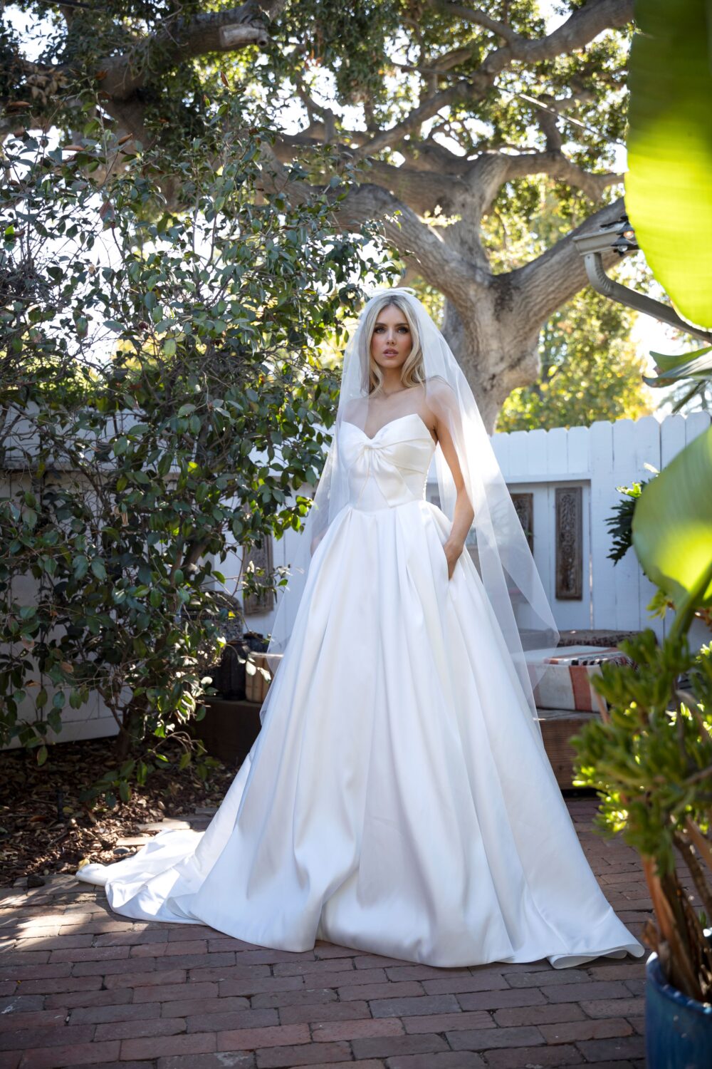 A bride in a strapless satin wedding dress with a sweetheart neckline and a bow at the waist stands on a brick patio wearing a long veil.