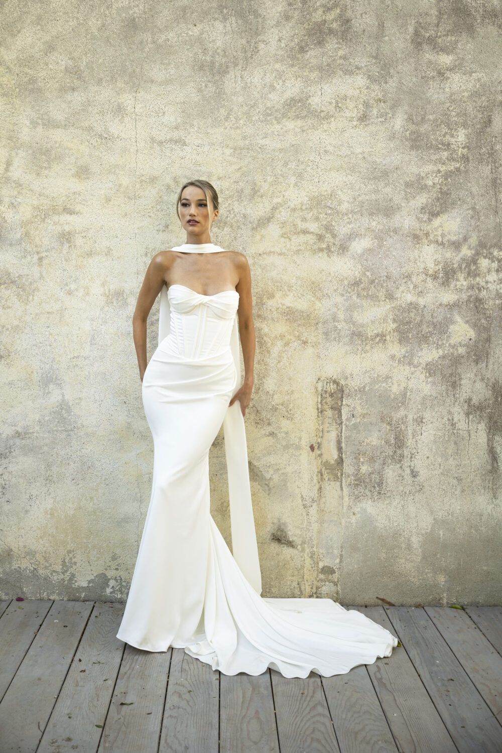 A strapless mermaid wedding dress with a ruched skirt and dramatic train is paired with a soft bridal scarf, modeled against a concrete wall.