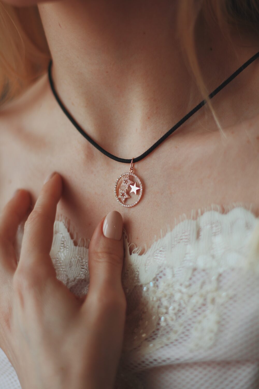 A close-up view of a delicate circular pendant with sparkling stars on a black cord necklace, resting on a lace-trimmed bridal neckline.