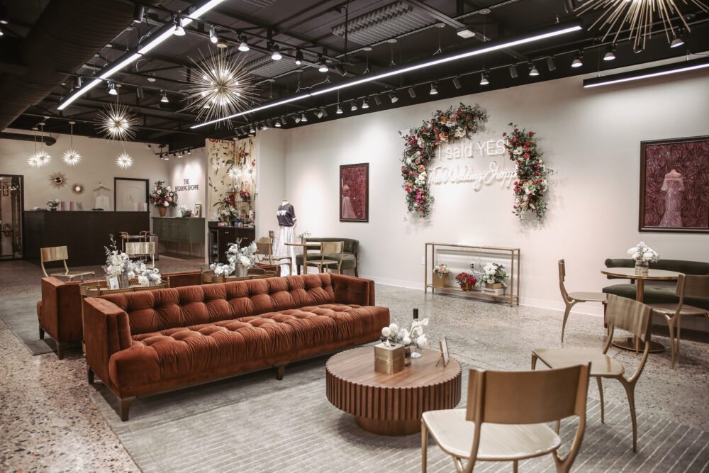 The Wedding Shoppe’s lobby features a rust-colored tufted couch, floral arch, and bright “I Said Yes” neon sign against a white wall.
