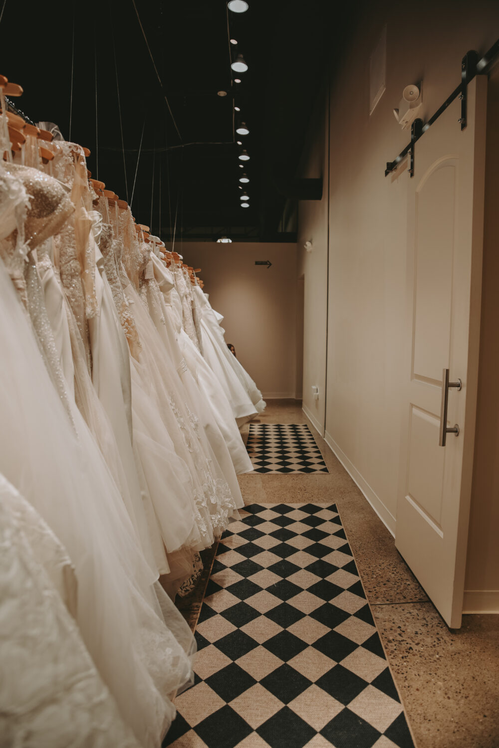 Another angle of the gown-lined hallway in a bridal shop, showing a partially open door and polished concrete floors with patterned rugs.