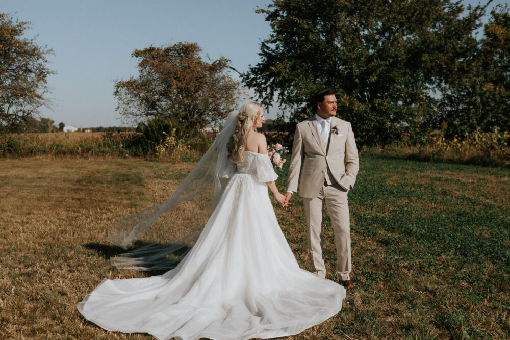 Bride and groom holding hands in a field during a daytime wedding, with the bride’s long train and veil trailing behind her.