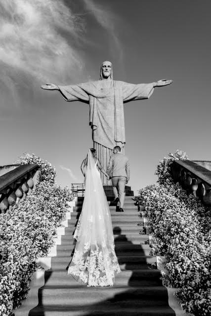 Bride and groom walking up the floral-lined steps toward the Christ the Redeemer statue in a dramatic black and white wedding photo.