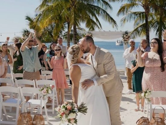 Couple kissing at the end of their beach wedding ceremony, surrounded by cheering guests under palm trees and string lights.